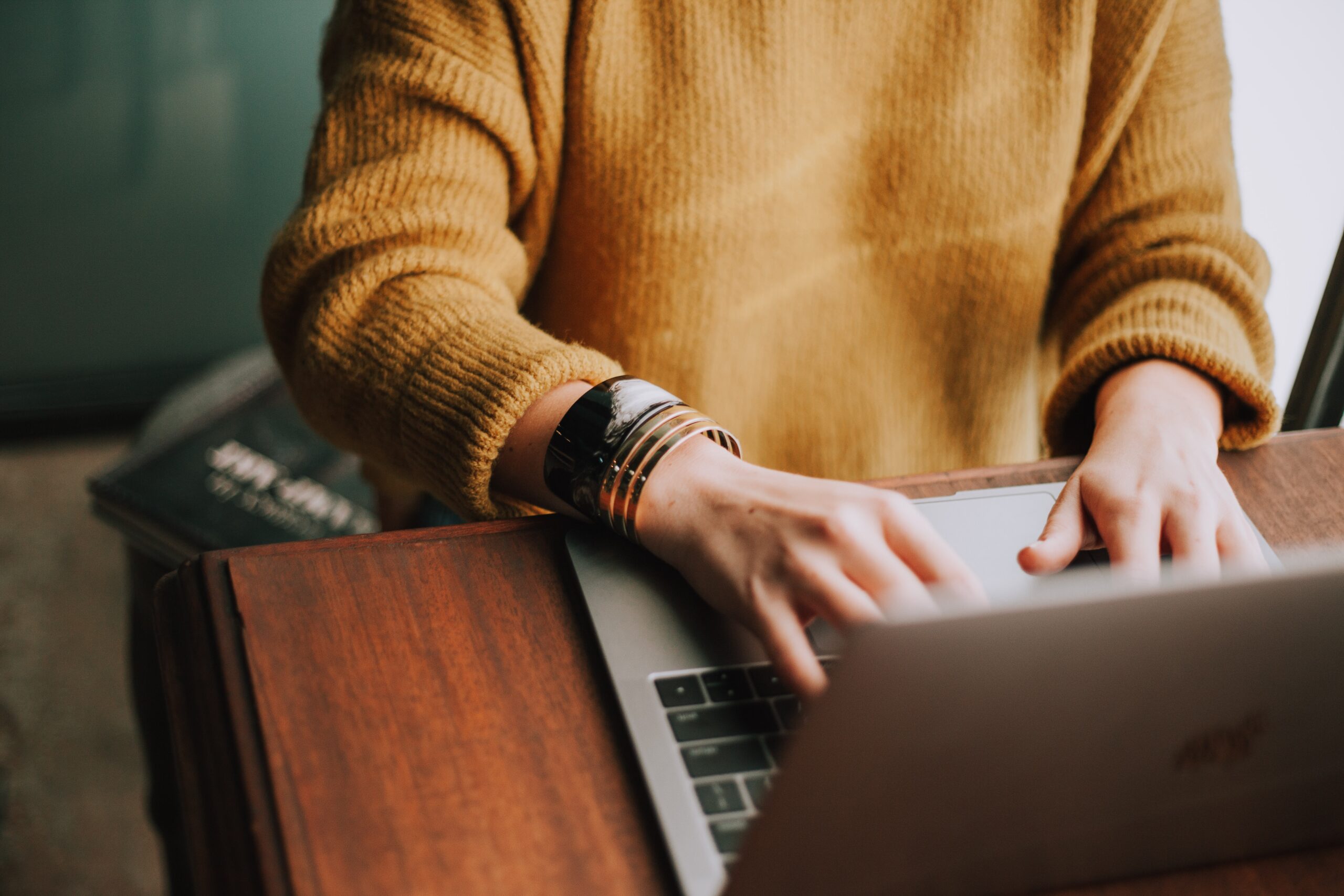 Zu sehen ist ein Oberkörper in einem gelben Pullover. Die Hände bewegen sich über die Laptop tastatur, welcher vor der Person auf einem braunen Tisch steht.